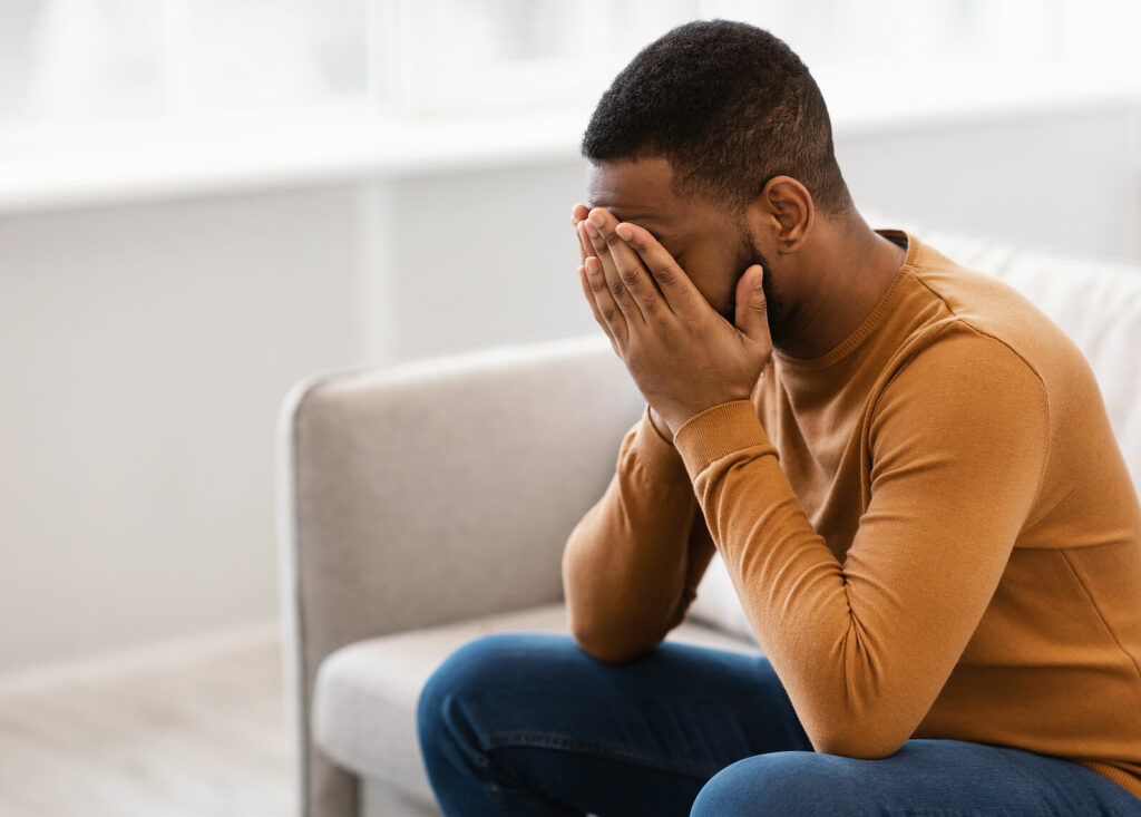 A man sits on the couch with his head in his hands as he struggles with depression. Therapy for depression in Washington, DC can help, reach out. 