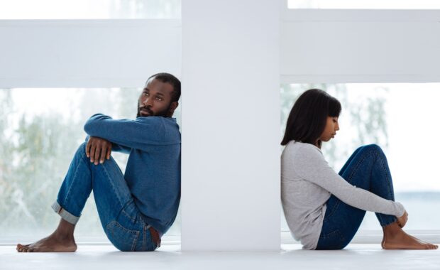 A man and woman sitting back-to-back on a white surface, both wearing denim and looking away from each other in contemplative poses, suggesting relationship distance or relationship anxiety