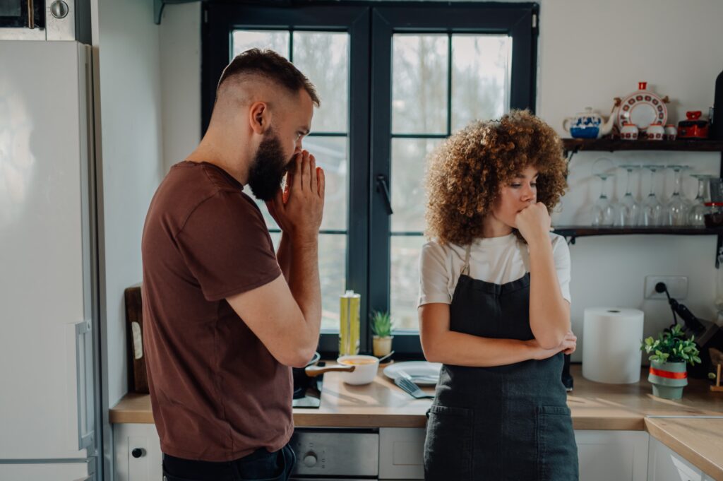 A bearded man in a brown shirt covering his face with his hand while a woman with curly hair in a white shirt and dark apron touches her chin thoughtfully, both appearing distressed during what seems to be a difficult conversation in a modern kitchen