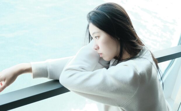 Woman leaning on railing gazing out window, reflecting on her mental health journey
