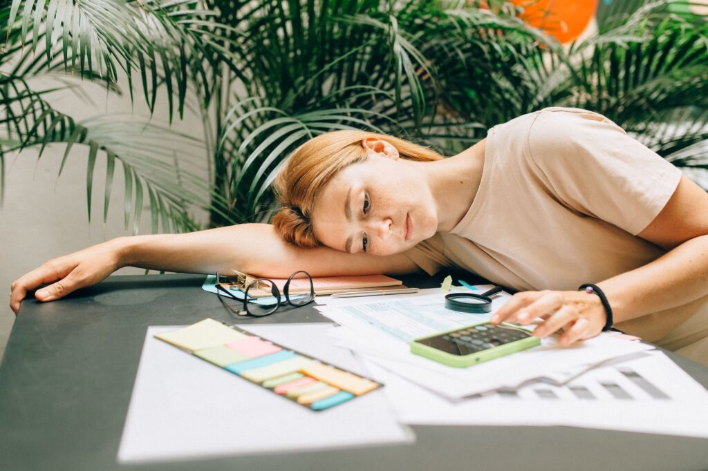 Woman resting her head on her arm at a cluttered desk, looking drained while surrounded by paperwork, sticky notes, and a phone