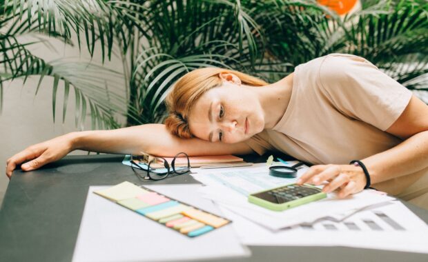 Woman resting her head on her arm at a cluttered desk, looking drained while surrounded by paperwork, sticky notes, and a phone